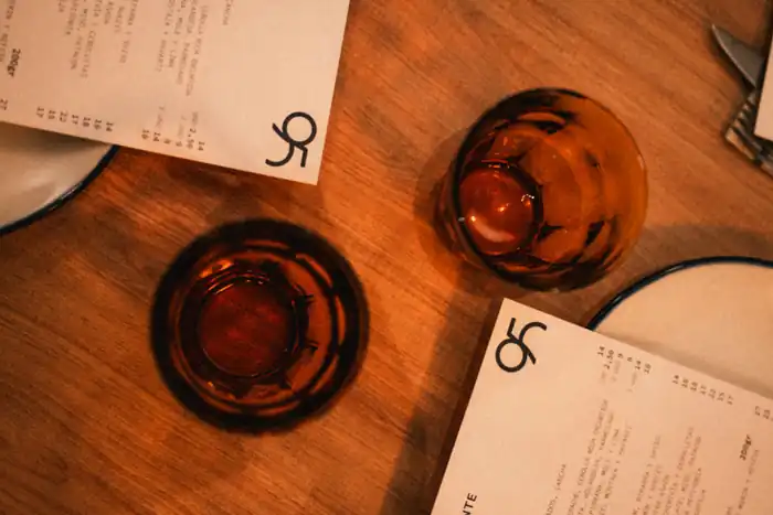 Dos vasos de vino ámbar sobre la mesa de madera, entre las cartas impresas de 95 y los platos de borde azul
