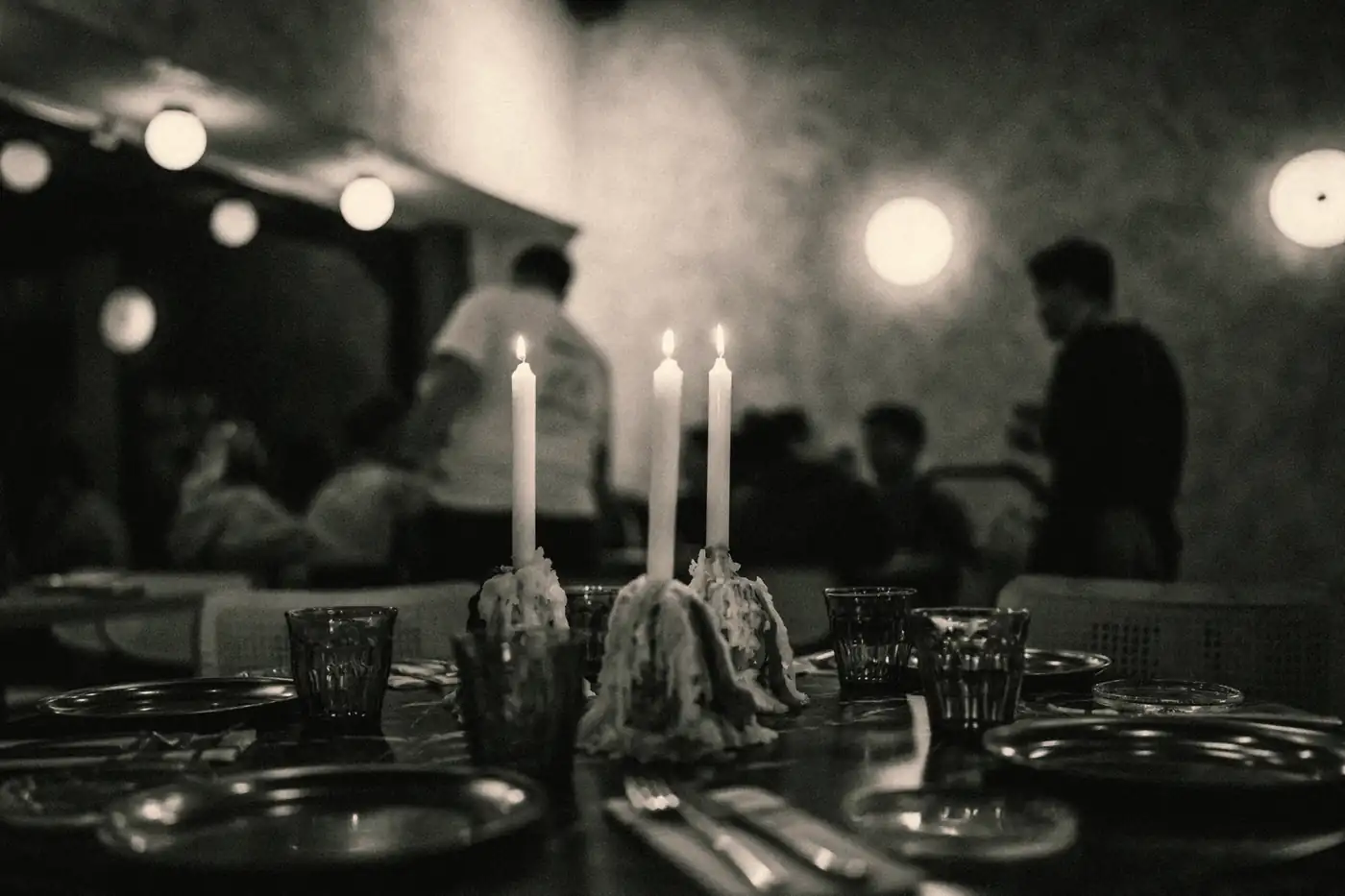 Tres velas encendidas sobre una mesa puesta de 95; detrás, Javier y Daniel atienden la sala llena (en blanco y negro)